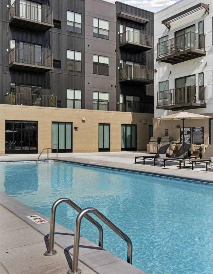 Outdoor swimming pool with metal ladder, lounge chairs, and modern apartment building in the background.
