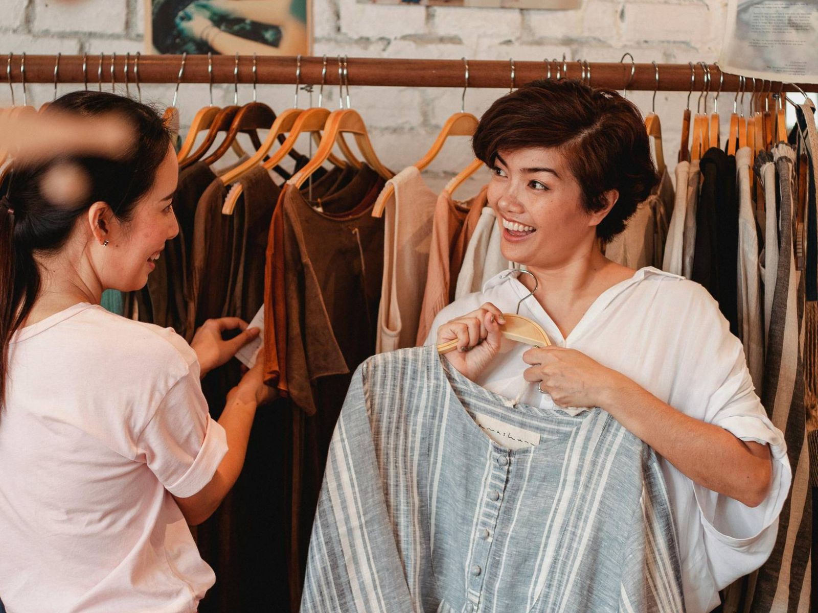 Two women smile and talk while shopping for clothes in a boutique with dresses hanging on racks.