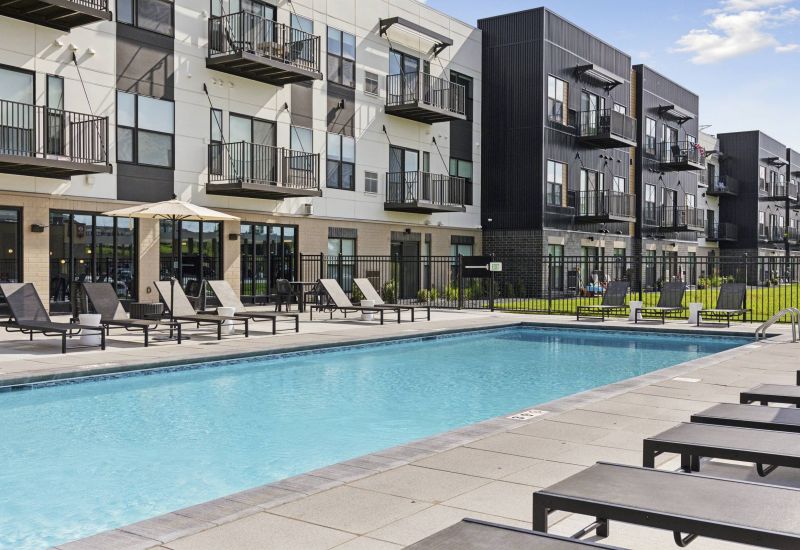 Outdoor apartment pool area with lounge chairs, umbrella tables, and modern building in the background.