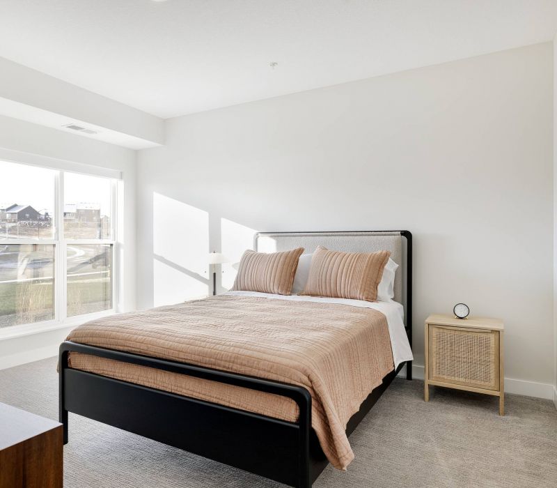 Bright bedroom with a bed, beige bedding, a nightstand, and large windows letting in natural light.