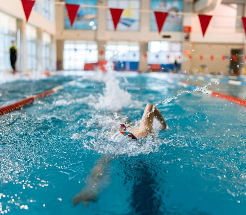 A swimmer in a pool swims backstroke, splashing water, with red lane dividers and flags overhead.