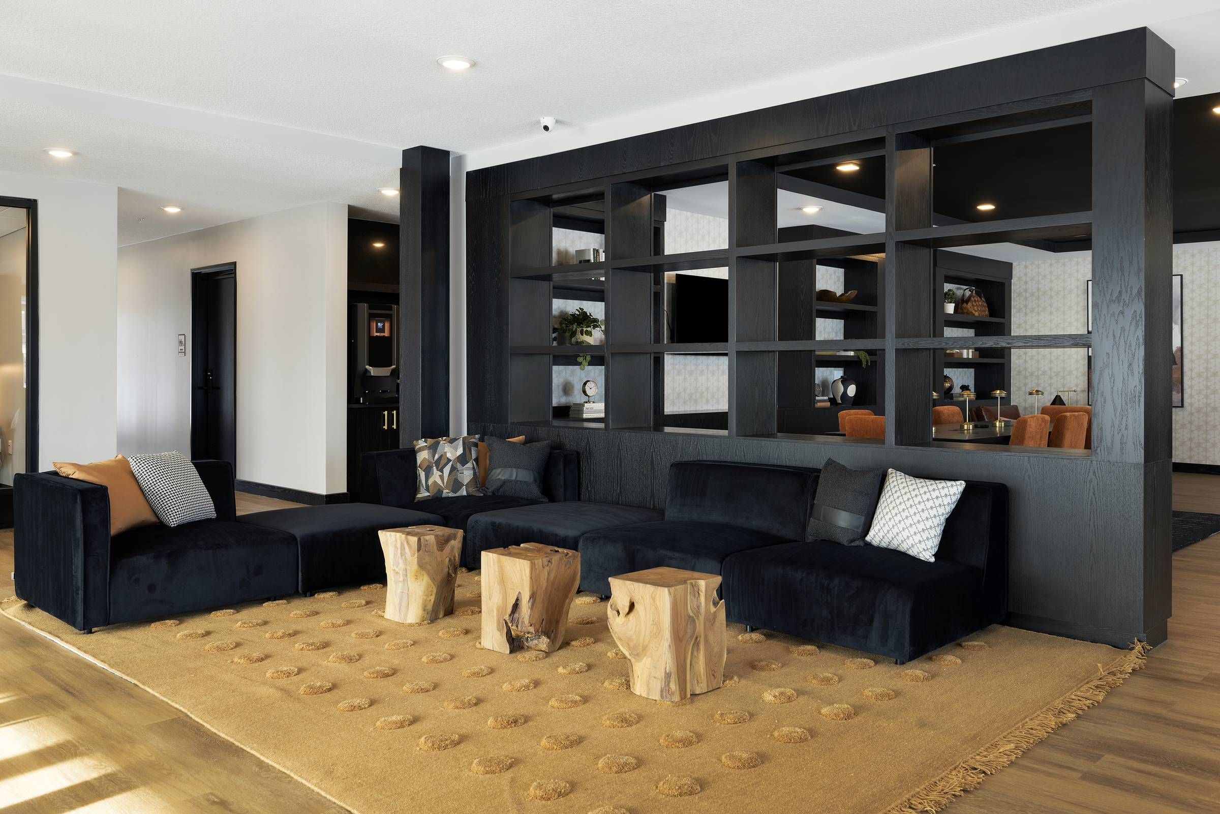 Modern lounge with black sectionals, wood stump tables, and a tan textured rug in front of an open black shelf.
