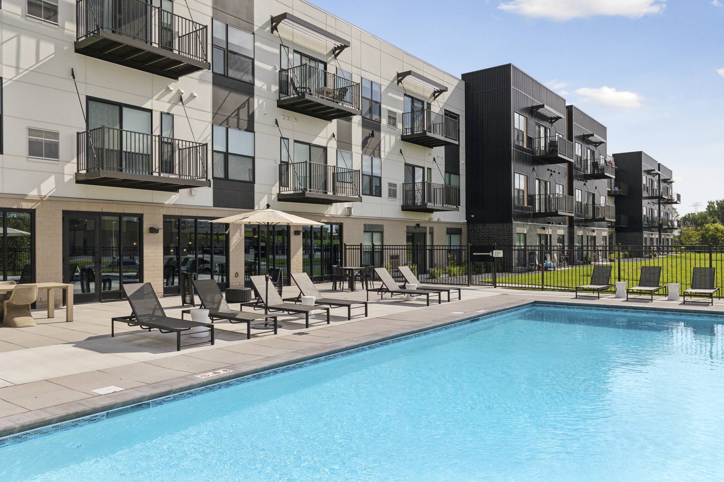 Modern apartment complex with balconies, poolside lounge chairs, and a clear blue swimming pool on a sunny day.