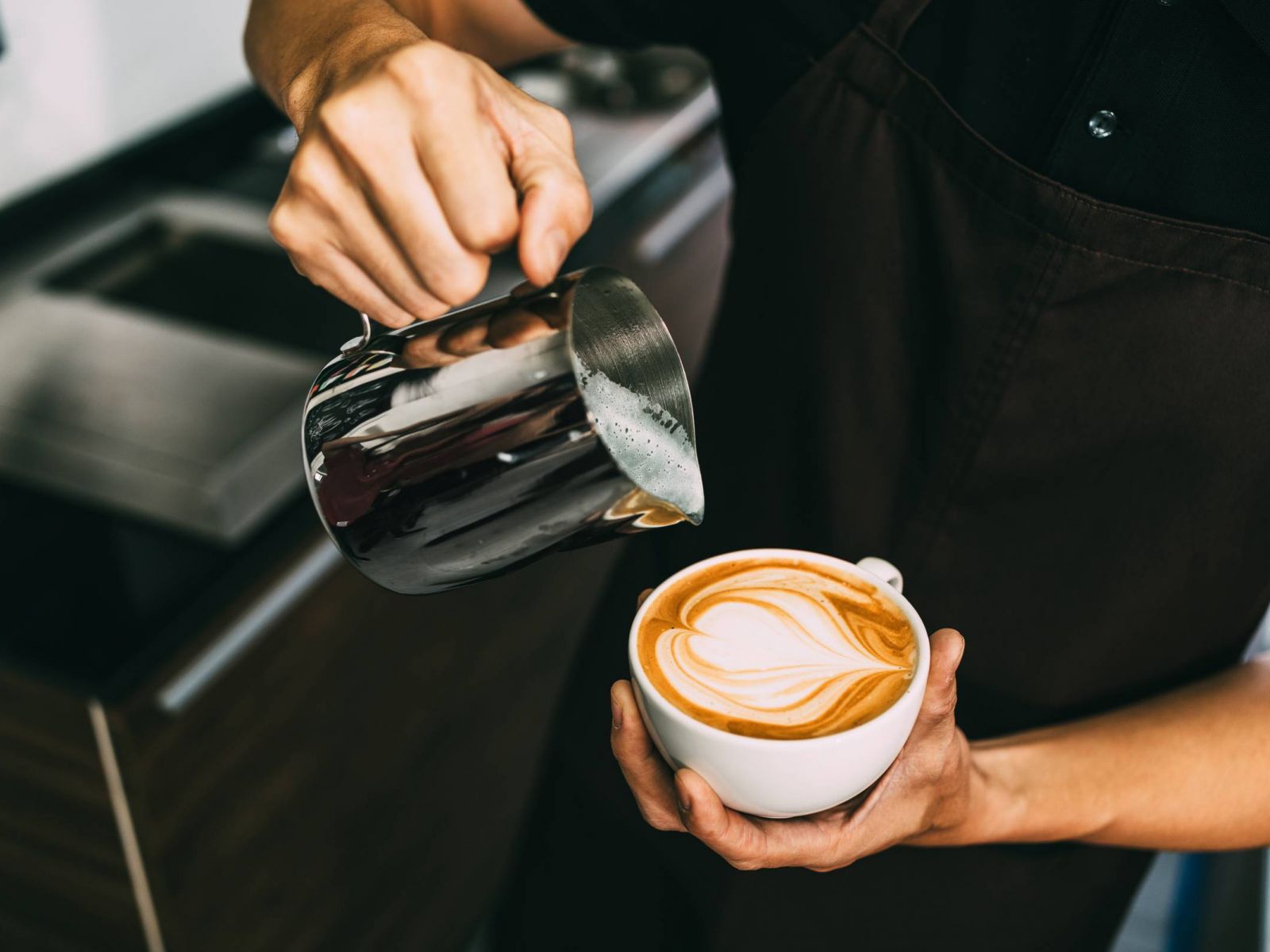 Barista pouring steamed milk into a cup of coffee, creating latte art in the shape of a heart.