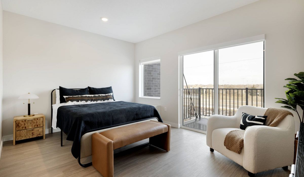 Modern bedroom with a bed, bench, armchair, nightstand, and balcony access through sliding glass doors.