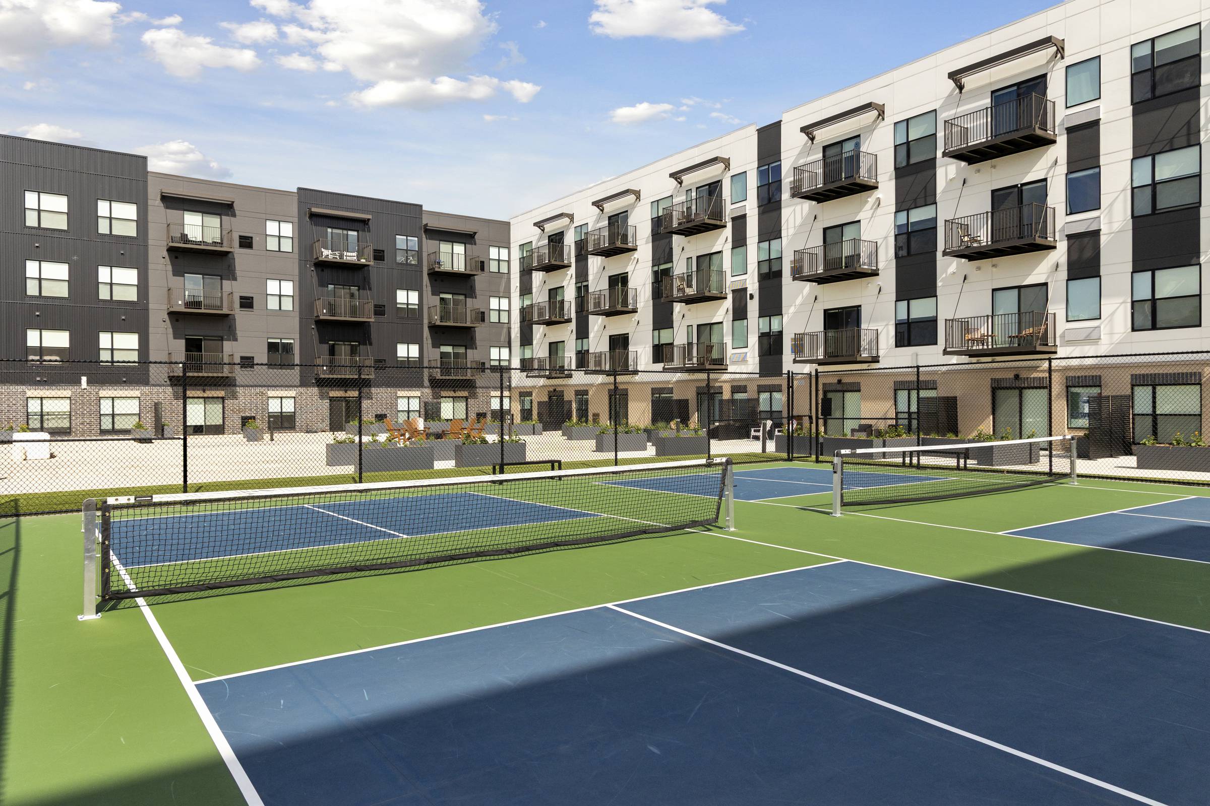 A modern apartment complex with balconies overlooks a blue and green outdoor pickleball court.