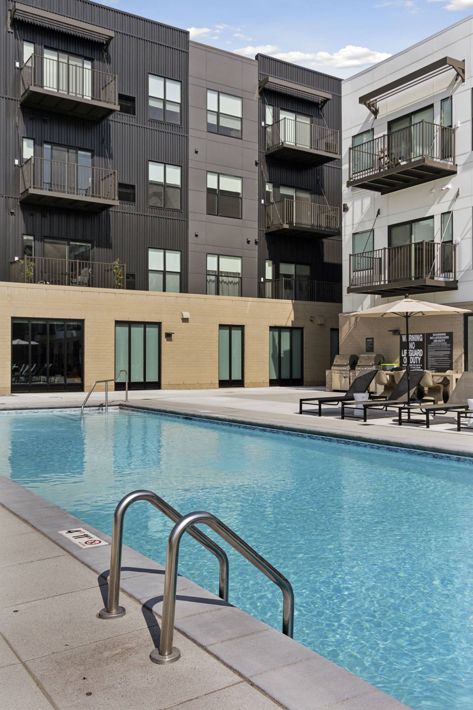 Outdoor swimming pool with metal ladder, lounge chairs, and modern apartment building in the background.