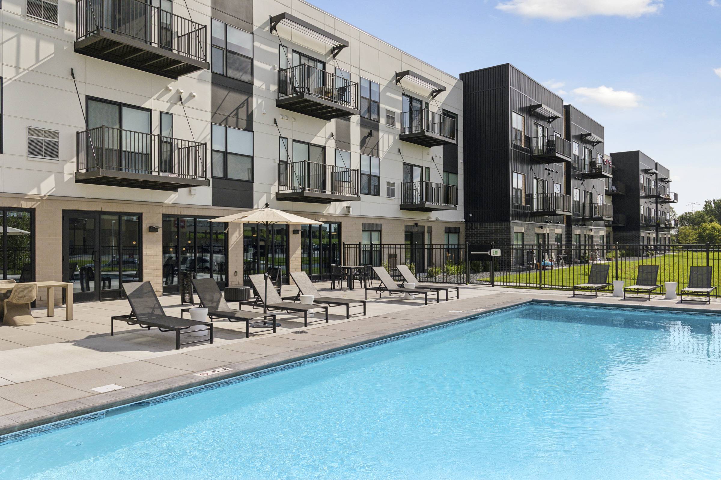 Modern apartment complex with balconies, poolside lounge chairs, and a clear blue swimming pool on a sunny day.