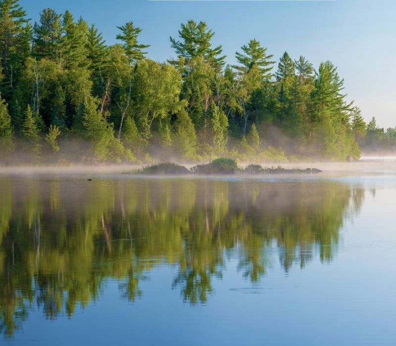 Calm lake with mist and reflections of green trees under a clear blue sky.
