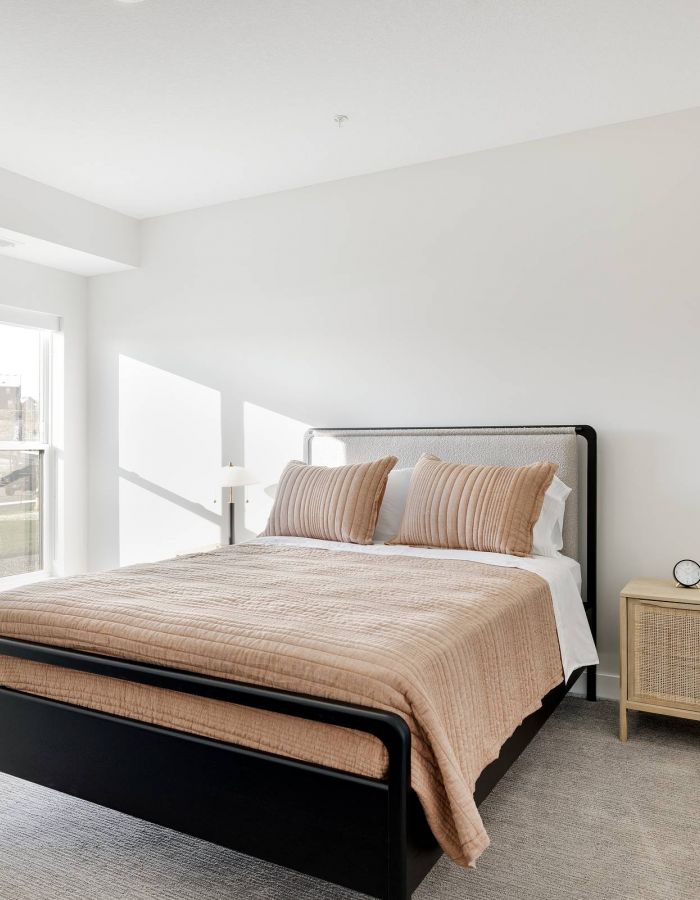 Bright bedroom with a bed, beige bedding, a nightstand, and large windows letting in natural light.
