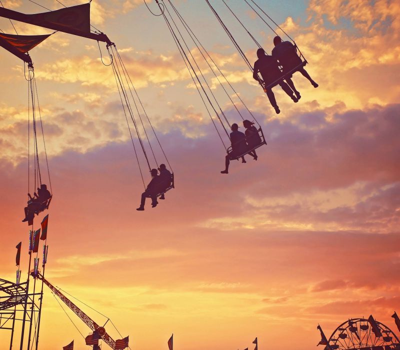 Silhouetted people ride swings at a carnival during a vibrant sunset with orange and purple clouds.