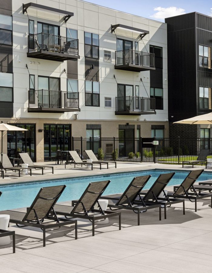 Outdoor swimming pool with lounge chairs beside a modern apartment building under a blue sky with scattered clouds.